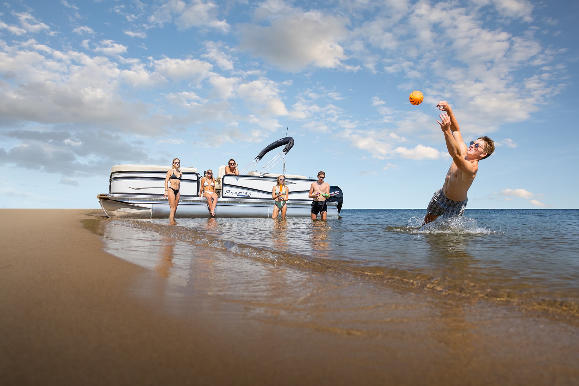 A group of young adults wearing swimsuits standing in front of a Premier tritoon that is pulled up onto a beach. A young man is diving for a thrown ball.