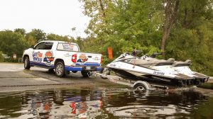 A Lake Fun Rentals pickup truck towing a trailer with a jet ski on it backing it up into the water.