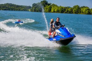 A man and woman riding a blue Yamaha jet ski. The woman is sitting backwards watching a young child who is being towed behind on an inflatable raft.