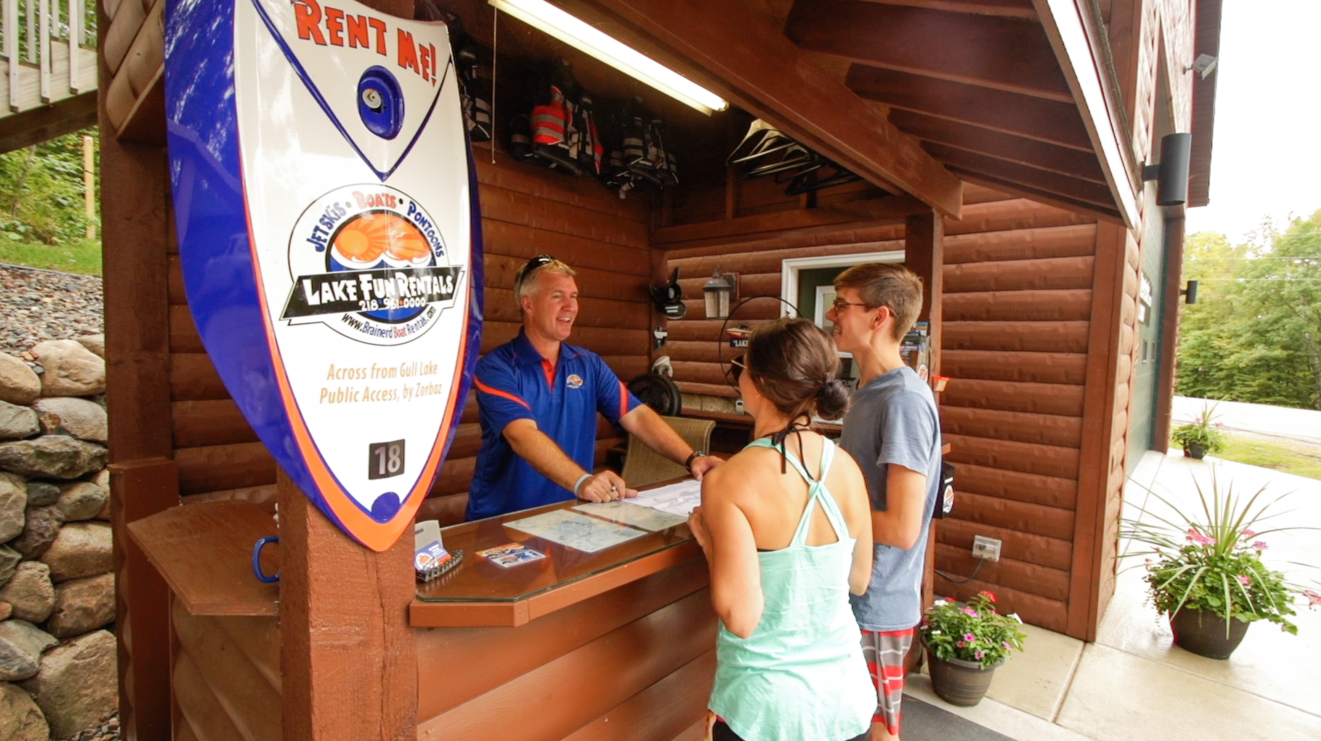A young man and woman standing in front of a Lake Fun Rentals desk while an employee helps them rent something.