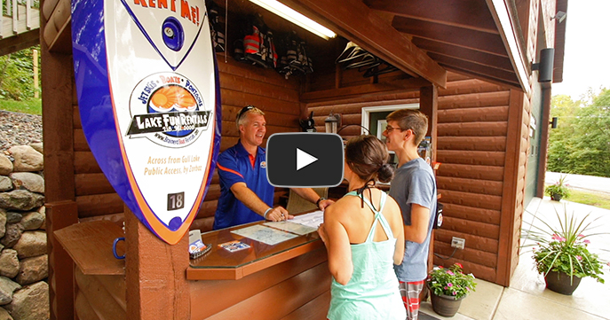 A video still image of a young man and woman standing in front of a Lake Fun Rentals desk while an employee helps them rent something.