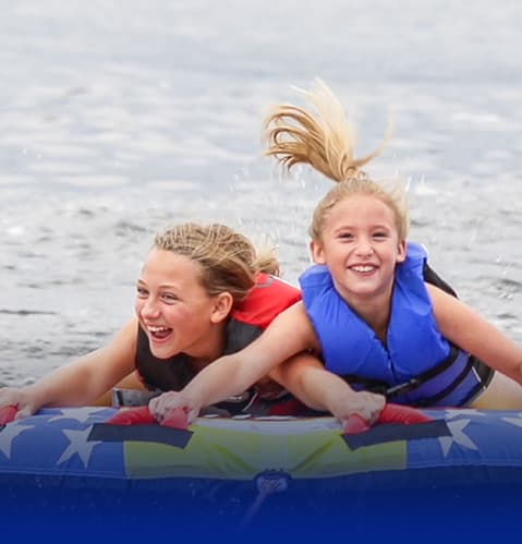 Two smiling young girls on an inflatable raft being towed by a boat.