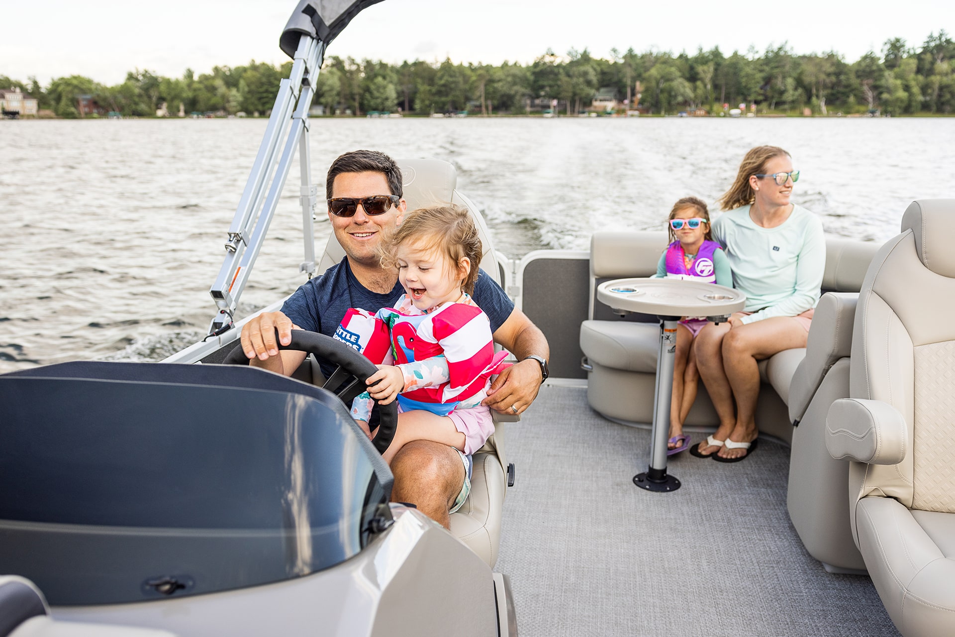 A head-on shot of a family driving a pontoon on a lake. The father is driving while one of the young girls is sitting on his lap. The mother and other young girl is sitting on seats in the back.