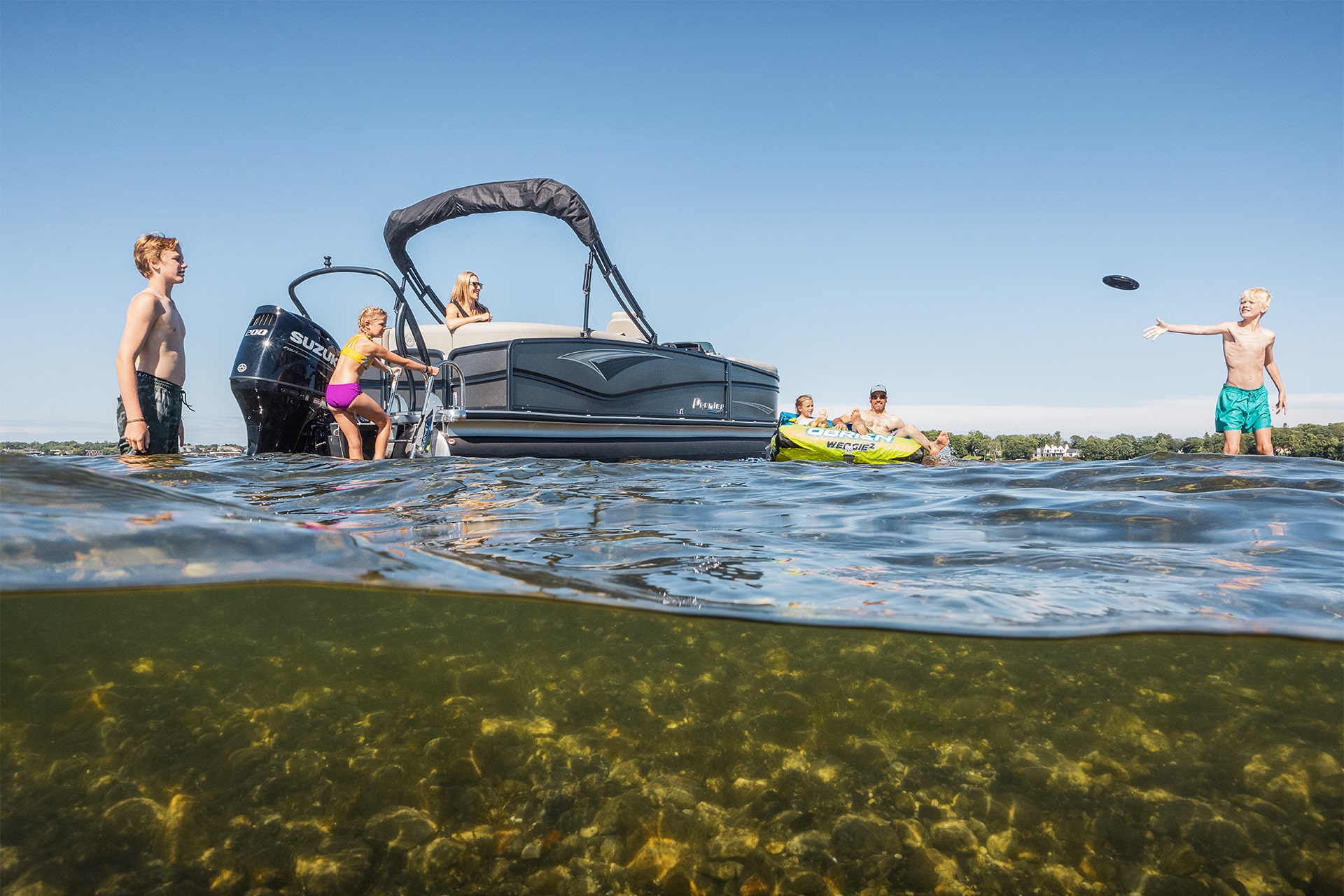 A family playing in a lake next to a Premier pontoon. A young boy is throwing a frisbee to another boy. A father and daughter are sitting on a raft. The mother is sitting on the pontoon while another daughter is climbing up a ladder on the back of the pontoon.