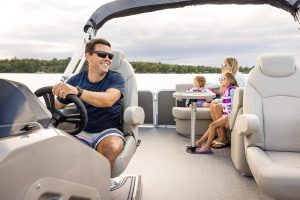 Father smiling while driving a pontoon while mother and daughters ride in the back.
