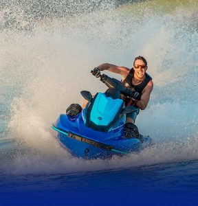 Smiling man with sunglasses on and wearing a lifevest riding a jet ski on a lake. He is turning the jet ski which is kicking up a big wave of water.