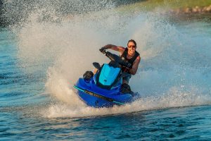 Smiling man with sunglasses on and wearing a lifevest riding a jet ski on a lake. He is turning the jet ski which is kicking up a big wave of water.